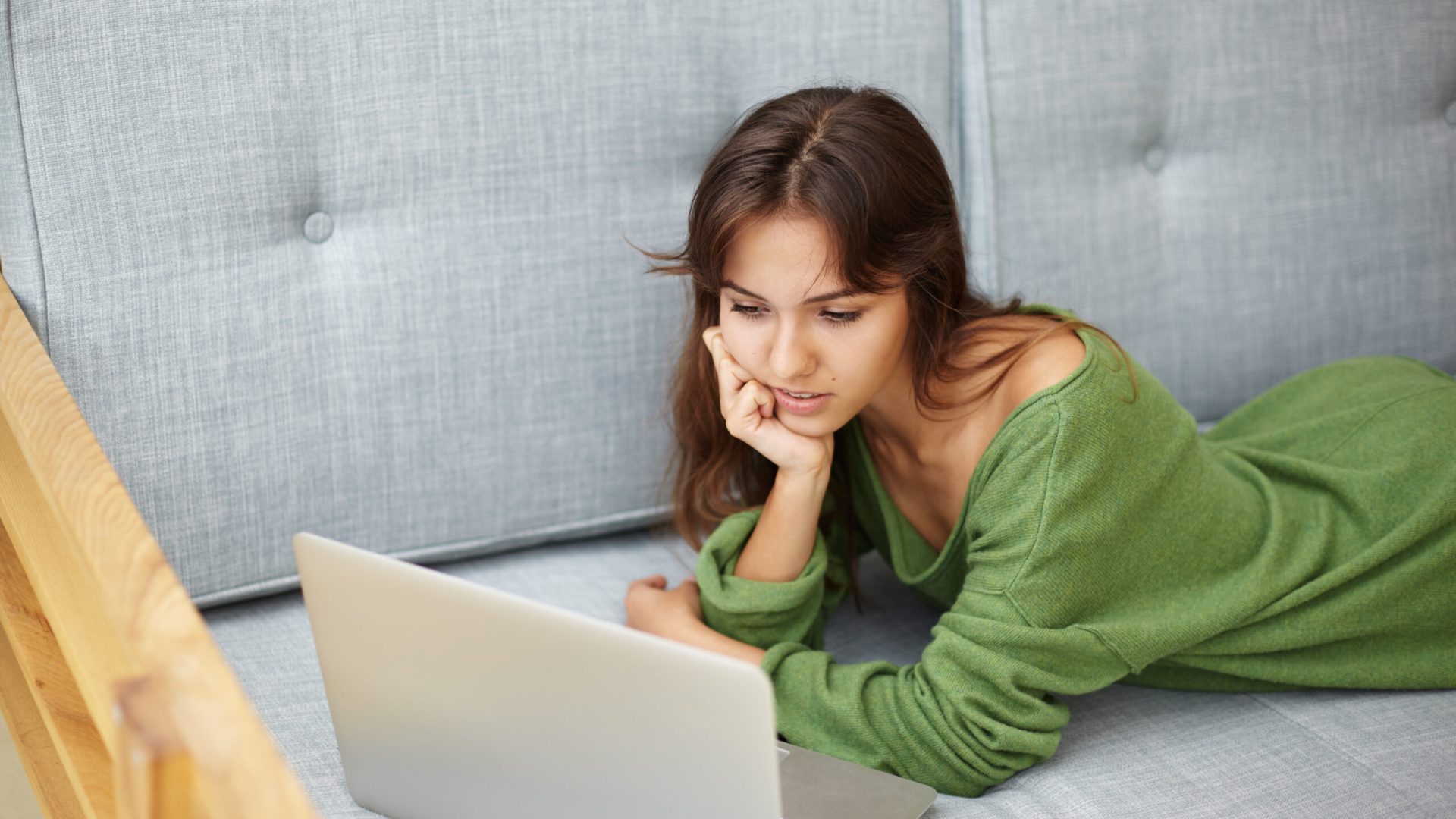 Attractive brunette young female surfing internet on generic notebook computer indoors, lying on grey sofa, booking plane tickets online, planning vacations. People and modern technologies concept