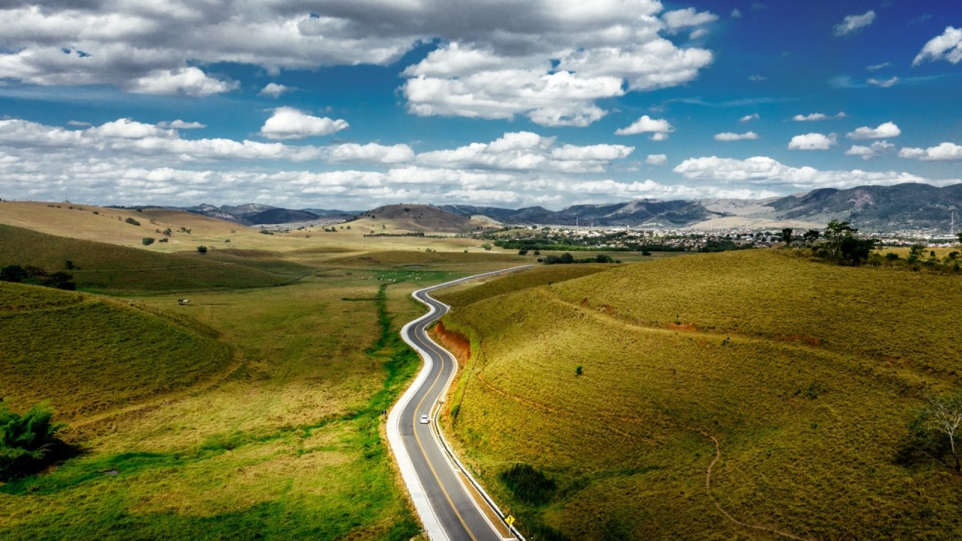 A road surrounded by hills covered in greenery with mountains under a cloudy sky on the background