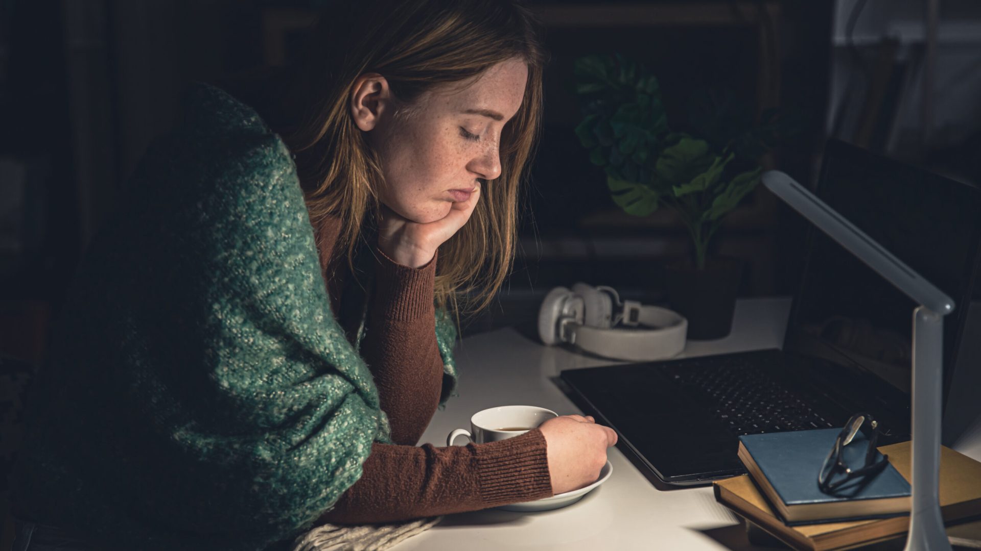 A tired young woman with a cup of coffee at the workplace late at night wants to sleep, insomnia, anxiety.